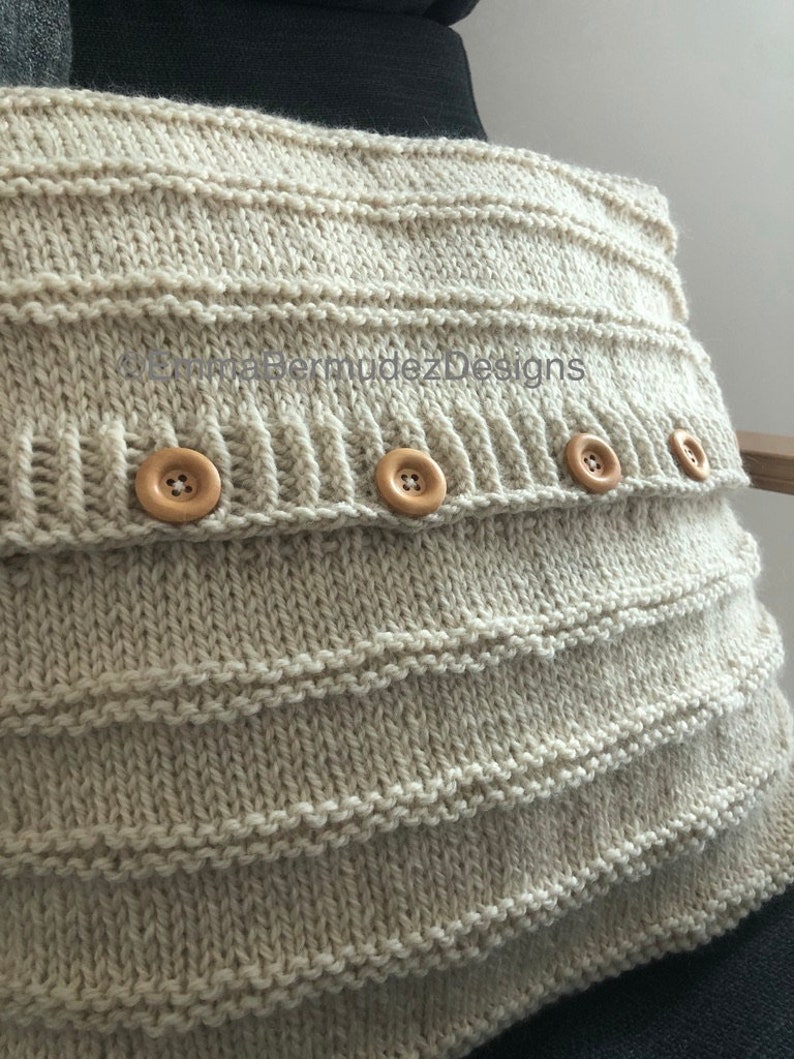 May include: A beige knitted pillow cover with horizontal ribbed detailing and four wooden buttons. The pillow cover is made of a soft, textured yarn and has a simple, classic design. The text "BermudezDesigns" is visible on the pillow cover.