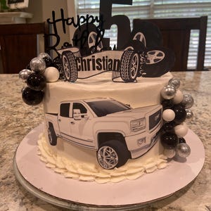 May include: A white birthday cake decorated with a white GMC truck, a black number 5, and a black cake topper that says "Happy Birthday Christian". The cake is adorned with black, silver, and white decorative balls.