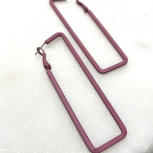 Pode incluir: Um par de brincos de argola retangulares em cor malva escura. Os brincos são feitos de metal e têm um design simples e moderno.