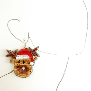 May include: A pair of beaded reindeer earrings with a red and white Santa hat. The reindeer have brown and tan beading and a white pearl nose.
