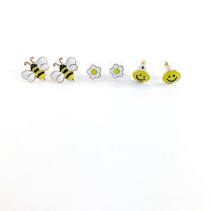 May include: A set of six gold-tone earrings featuring a bee, flower, and smiley face design. The bee earrings are yellow and black, the flower earrings are white, and the smiley face earrings are yellow.