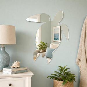 May include: A sea turtle-shaped wall mirror with a reflective surface that captures the surrounding room, including a window, a framed picture, and a potted plant. The mirror features a distinct silhouette of a swimming sea turtle.