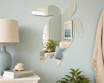 May include: A sea turtle-shaped wall mirror with a reflective surface that captures the surrounding room, including a window, a framed picture, and a potted plant. The mirror features a distinct silhouette of a swimming sea turtle.