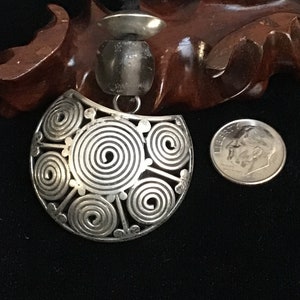 May include: A silver pendant necklace with a large, circular, openwork design featuring multiple concentric circles. The pendant is attached to a black cord.