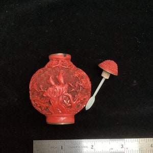Pode incluir: Uma pequena garrafa de rapé de cinábrio vermelho esculpida com um design detalhado de um pássaro e galhos. A garrafa tem uma borda de latão e uma pequena rolha vermelha em forma de cogumelo com uma haste de metal.