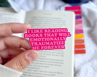 Puede incluir: Una pegatina rosa con el texto "I LIKE READING BOOKS THAT WILL EMOTIONALLY TRAUMATIZE ME FOREVER" se sostiene frente a un libro abierto. La pegatina está en una fuente de letras de burbujas. Una pila de notas adhesivas de colores y una vela azul oscuro están en el fondo.