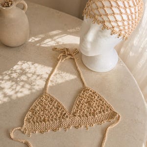 May include: A beige crochet halter top and a matching crochet headpiece, both featuring an open net pattern and small dangling metallic charms along the edges. The halter top has thin tie straps, and the headpiece is displayed on a white mannequin head.