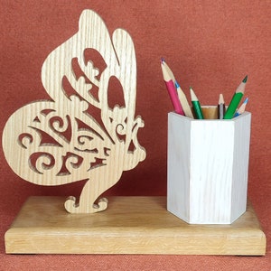 Peut inclure: Un porte-crayons en bois en forme de papillon avec un pot à crayons hexagonal blanc. Le papillon est finement sculpté avec un motif floral et la base est en bois naturel.