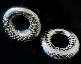 Op de afbeelding: Een paar cirkelvormige zilverkleurige oorringen met een textuur van een ruitvormig rasterpatroon op de onderste helft van elke ring. Het bovenste gedeelte van elke ring is glad en gepolijst, met een zichtbaar scharnier voor sluiting.