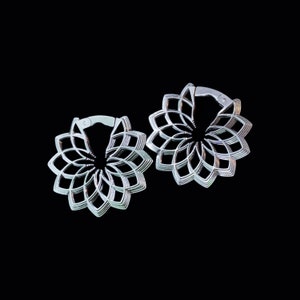 Puede incluir: Un par de pendientes de aro plateados con un diseño de flor geométrico. Los pendientes están hechos de metal y tienen un acabado brillante.