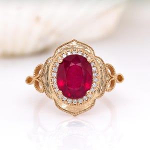Könnte beinhalten: Ein goldener Ring mit einem ovalen, tiefroten Edelstein, umgeben von kleinen, klaren Steinen. Der Ring hat ein aufwendiges, filigranes Design um den zentralen Stein und am Band, im Vintage-Stil.