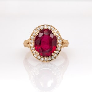 May include: A gold ring with a large, oval ruby encircled by a halo of small diamonds. The ring's band has a detailed, textured pattern. The ruby is a rich, deep red, and the diamonds glisten against the gold setting.