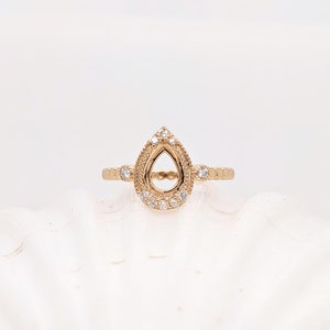 May include: A gold ring with a pear-shaped setting surrounded by small diamonds. The ring is designed to hold a larger pear-shaped gemstone.