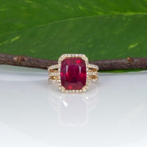 May include: A gold ring featuring a large, rectangular, cushion-cut deep red gemstone at the center. The stone is surrounded by a halo of small, clear, round-cut stones. The band is split into three rows, each encrusted with small, clear stones.