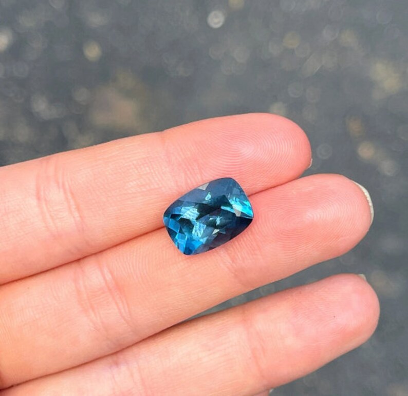 May include: A deep blue gemstone with a cushion cut, displayed on a finger.