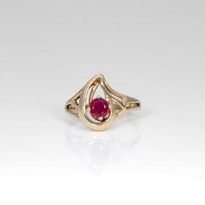 May include: A gold ring featuring a round, vibrant red gemstone set within a swirling, open-work metal design. The band curves gracefully around the central stone, creating a fluid, asymmetrical shape that highlights the polished gold finish.