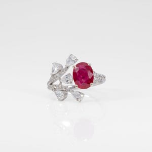 May include: A ring featuring a central oval-cut red ruby gemstone set in a silver-toned band. The band is adorned with multiple small, clear, pear-shaped and round-cut stones arranged in a branch-like pattern extending from the ruby.