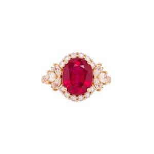 May include: A gold ring featuring a large, oval-cut ruby center stone surrounded by a halo of small, round diamonds. Additional pear-shaped diamonds flank the ruby, creating a cluster design. The ring is set against a white background.