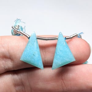 May include: Two light blue triangular gemstone beads strung on a silver wire with blue thread tassels on each end.