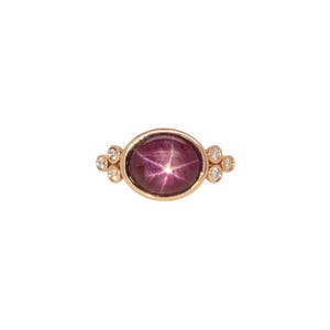 May include: A gold ring featuring a large, oval, purplish-red gemstone with a star-like pattern. Small, round, clear gemstones are set on either side of the main stone. The ring has a simple, elegant design.