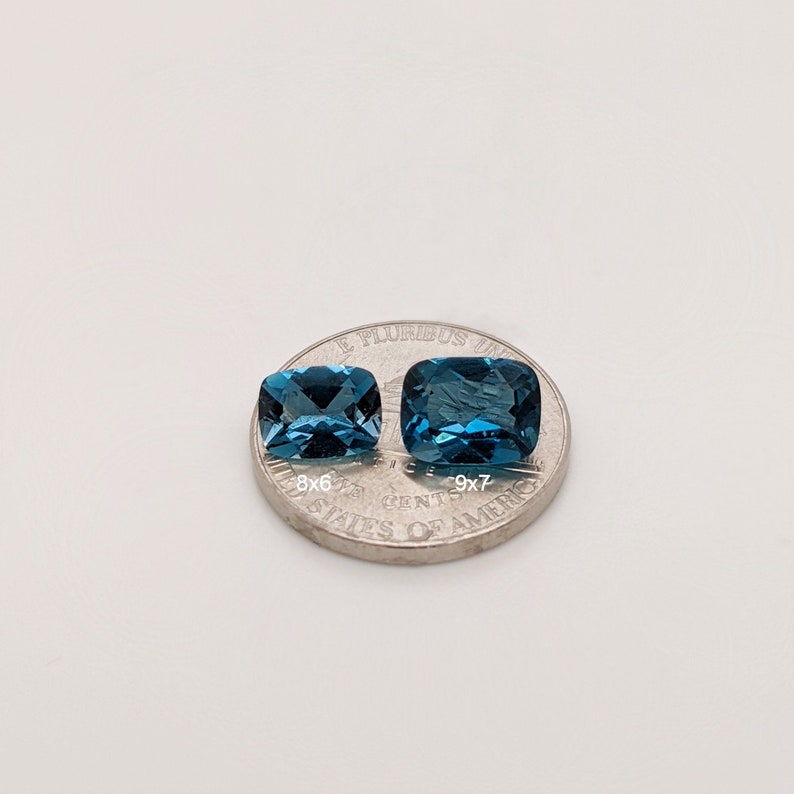 May include: Two blue topaz gemstones, one slightly larger than the other, are displayed on a US quarter. The smaller stone measures 8x6mm and the larger stone measures 9x7mm.