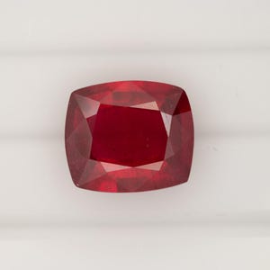 May include: A cushion-cut ruby gemstone with a deep, saturated red color. The stone features a faceted surface that reflects light, creating distinct geometric patterns across its top. The gemstone has a rectangular shape with rounded corners.