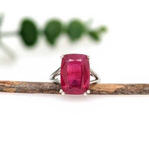 May include: A silver ring featuring a large, rectangular, ruby-red gemstone. The ring's band splits into a double-band design on each side of the gemstone. The ring is set against a neutral background with a wooden stick and greenery.