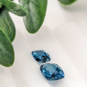 May include: Two cushion-cut blue gemstones on a white surface. The gemstones are a deep blue colour and have a slight sparkle.
