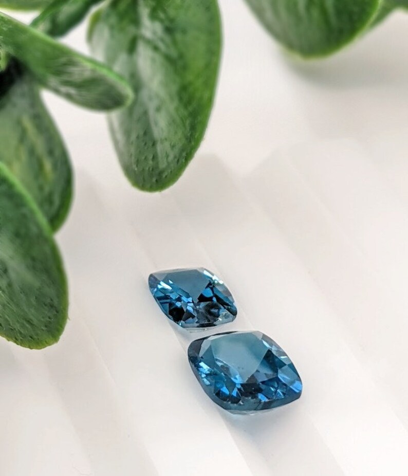 May include: Two cushion-cut blue gemstones on a white surface. The gemstones are a deep blue colour and have a slight sparkle.