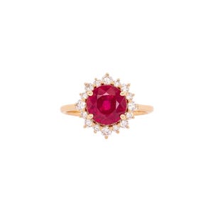 May include: A gold ring with a large, round, deep red gemstone encircled by smaller, clear gemstones. The ring has a halo design, with the central stone as the focal point, set against a white background.