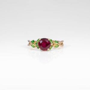 May include: A gold ring featuring a round, deep red center stone held by four prongs. The band is designed with a twisted vine motif, accented by small, marquise-cut green stones set along the sides, creating a botanical appearance.