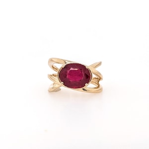 May include: A gold ring with a large, oval-shaped ruby gemstone set in the center. The ring band is made of three thin, intertwined bands.