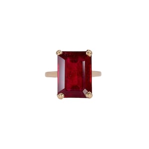 May include: A gold ring featuring a large, rectangular, emerald-cut deep red gemstone set in a four-prong mounting. The stone displays a rich, saturated crimson hue with visible internal facets, held securely by polished gold prongs on a simple band.