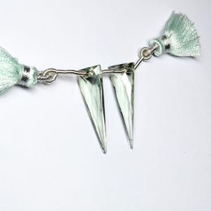 May include: Two light green faceted crystal beads with silver wire loops. The beads are shaped like a triangle with a pointed tip. The beads are attached to a silver wire with two light green tassels.