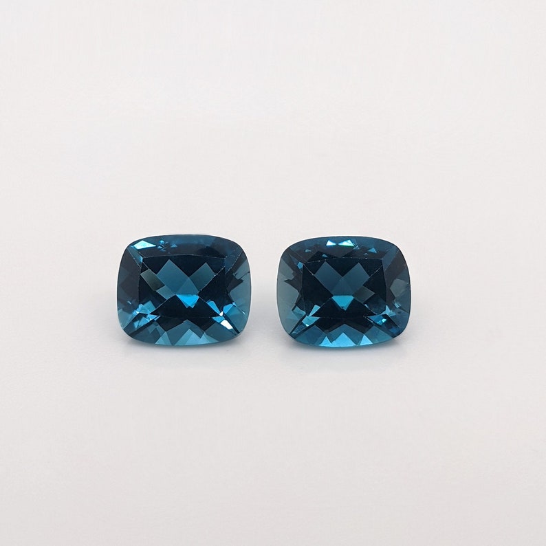 May include: Two cushion-cut, deep blue topaz gemstones. The gemstones are faceted and have a brilliant shine.