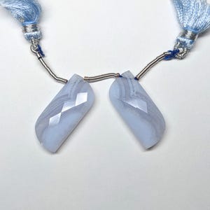 May include: Two light blue gemstone pendants with a faceted design, connected by silver wire and light blue tassels. The pendants have a rectangular shape with angled edges. The tassels are made of a soft, light blue material.