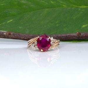 May include: A gold ring featuring a large, oval-cut, deep red gemstone at the center, flanked by three small, clear, round-cut stones on each side. The band has a polished gold finish with a ribbed, layered design extending from the center setting.