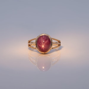 May include: A gold ring featuring a central oval cabochon-cut star ruby with a visible six-rayed star effect. The band is split into three distinct sections, with the outer two featuring a twisted rope texture and the center set with small diamonds.