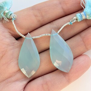 May include: Two light blue teardrop-shaped gemstones with faceted surfaces. Each gem is strung on a silver wire and adorned with a light blue tassel. The gemstones are held in a hand against a white background.