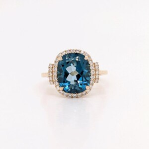 May include: A gold ring featuring a large, cushion-cut blue topaz gemstone encircled by a halo of smaller diamonds.