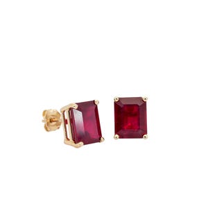 May include: A pair of gold stud earrings featuring rectangular, deep red gemstones. The earrings have a classic design with a gold setting that complements the vibrant color of the stones. The studs are displayed against a white background.