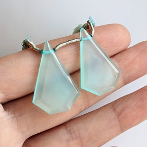 May include: Two light blue faceted gemstone pendants with silver wire loops. The pendants are held in a hand.