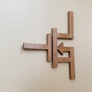 Può includere: Un'opera d'arte in legno montata a parete realizzata con più pezzi di legno disposti in un motivo geometrico. Il legno è di colore marrone chiaro e i pezzi sono disposti per formare una freccia che punta a sinistra.