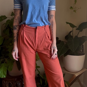 May include: A light blue t-shirt with white stripes on the sleeves and a white collar, paired with high-waisted, wide-leg coral pants. The shirt has a small logo on the chest. The pants have pleats at the waist.
