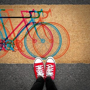 A pair of red and white sneakers stands on a doormat with a colorful bicycle design.