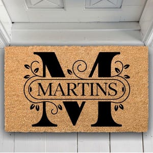 May include: A rectangular coir doormat featuring a black monogram "M" and the name "MARTINS" in an oval. The design includes floral elements. The mat is positioned in front of a white door.