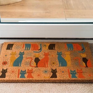 A colorful, patterned door mat with various cat illustrations on it, placed on a carpeted floor near a door.