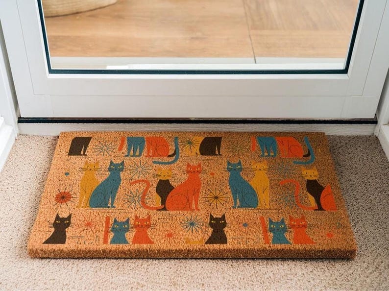 A colorful, patterned door mat with various cat illustrations on it, placed on a carpeted floor near a door.