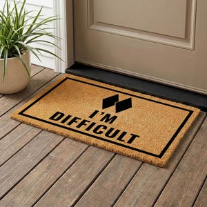 May include: A rectangular doormat made of natural coir fibre. The mat features a black border and the phrase "I'M DIFFICULT" in bold, black letters, along with two black diamond shapes. The mat is placed on a wooden porch.