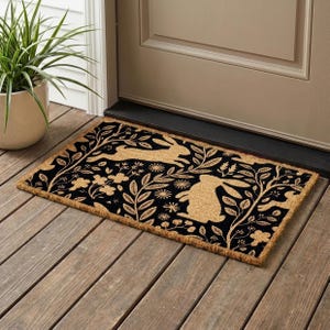 Peut inclure: Un paillasson rectangulaire en fibre de coco avec un fond noir et un motif en fibres naturelles beige représentant des lapins en course, des branches feuillues et des petites fleurs. Le design est symétrique sur toute la surface.